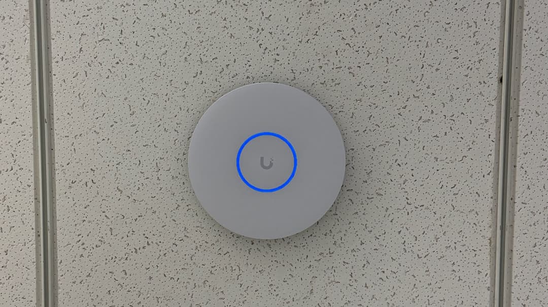 Round UniFi access point installed on an open industrial ceiling near overhead LED linear lights